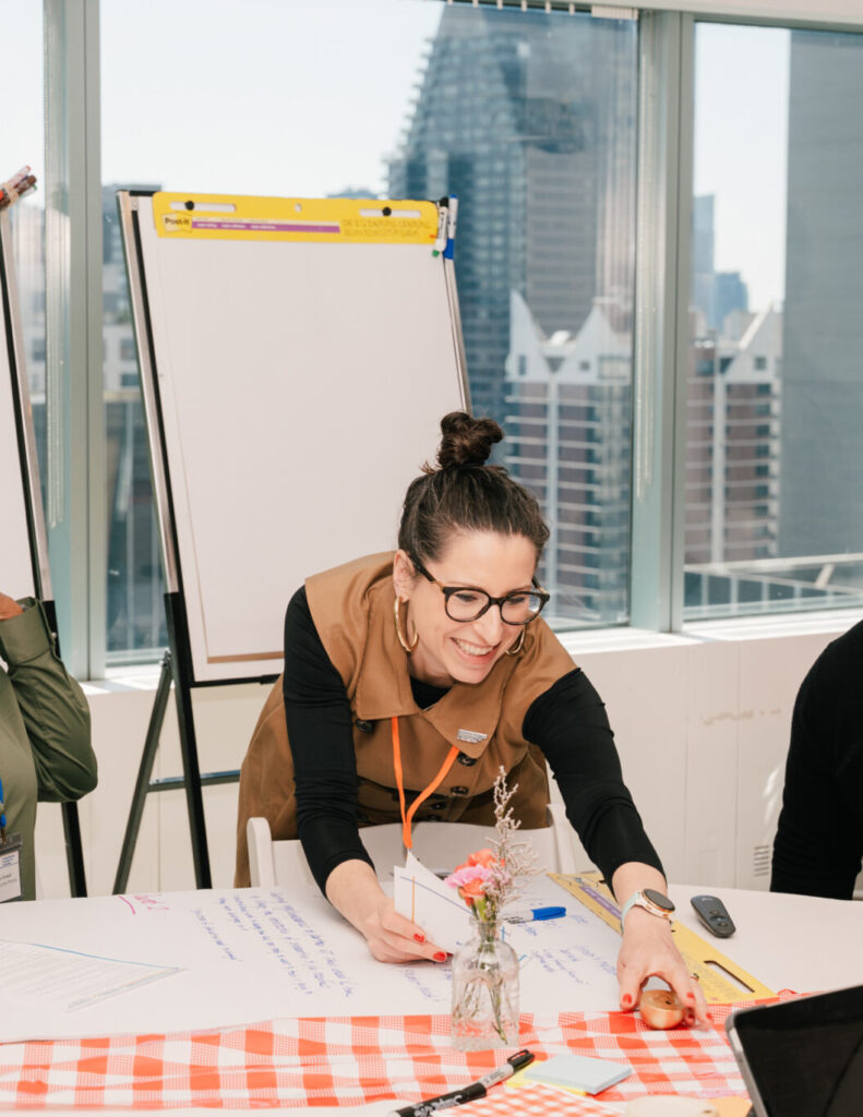Angela leans over a table covered in creative work, with flip charts and the New York skyline in the background